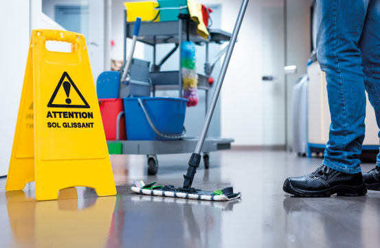 Reinigungskraft kehrt Flur, im Hintergrund Reinigungswagen und Warnschild Achtung nass